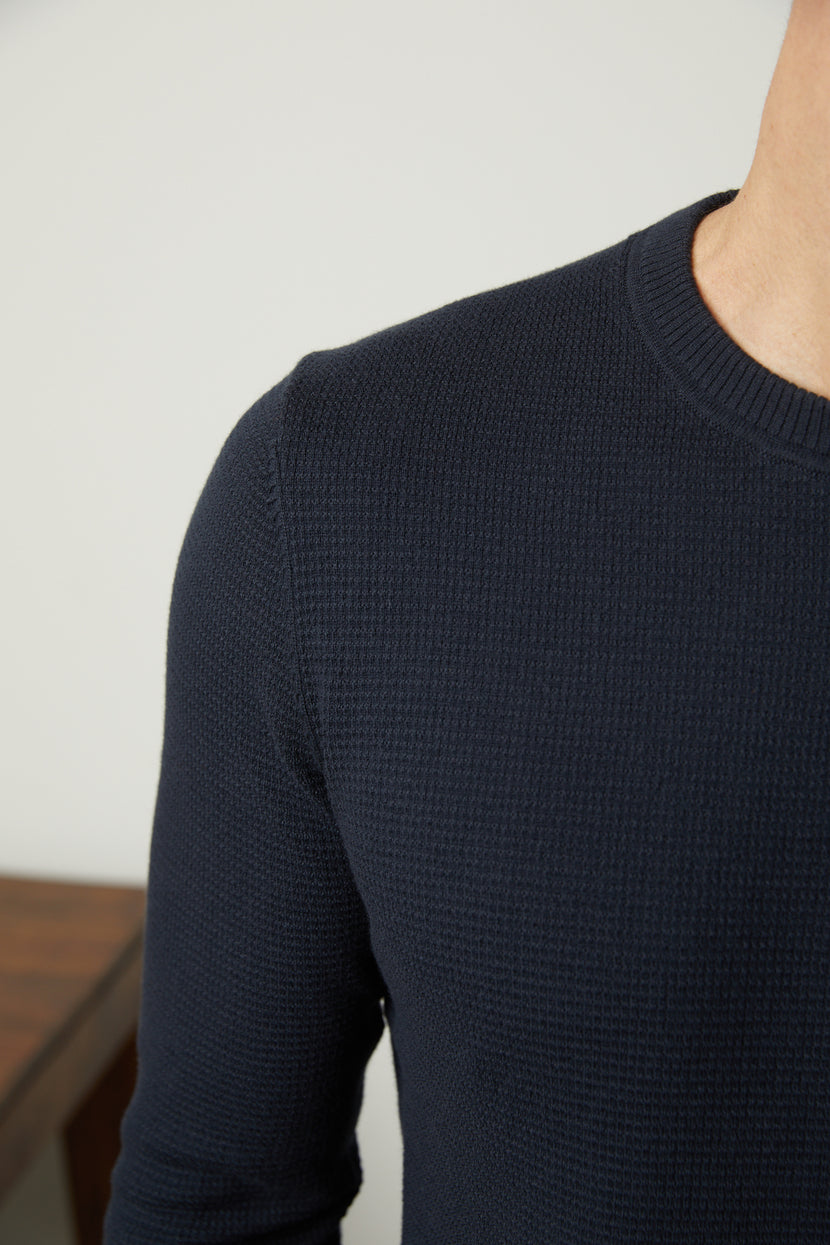 Person wearing the WALTER SWEATER by Velvet by Graham & Spencer, a black, knit long-sleeve shirt with a subtle thermal design, shown from the upper chest to shoulder area, standing in front of a white wall and a wooden surface.