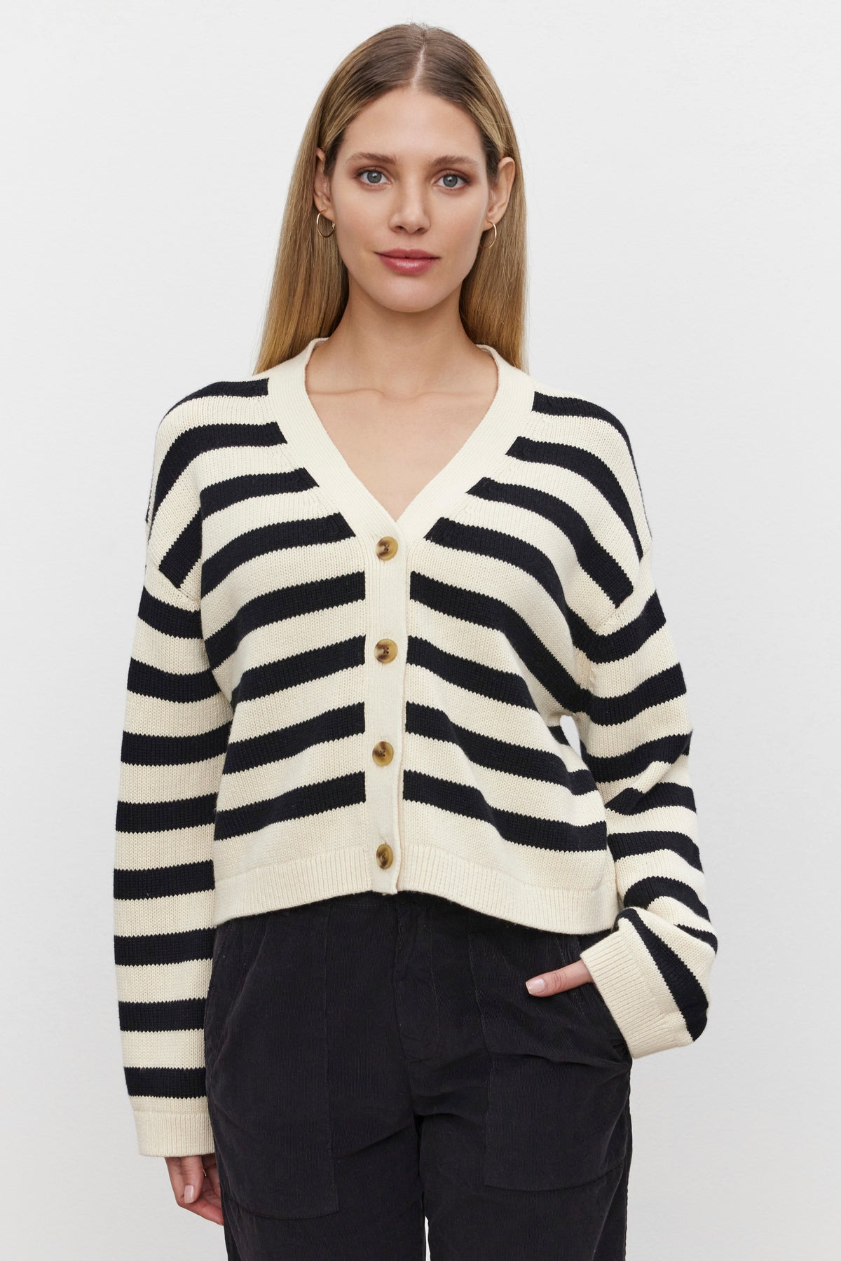   A woman wearing the ELODIE CARDIGAN by Velvet by Graham & Spencer, featuring black and white striped details and gold buttons, stands against a plain background with one hand in her pant pocket. 