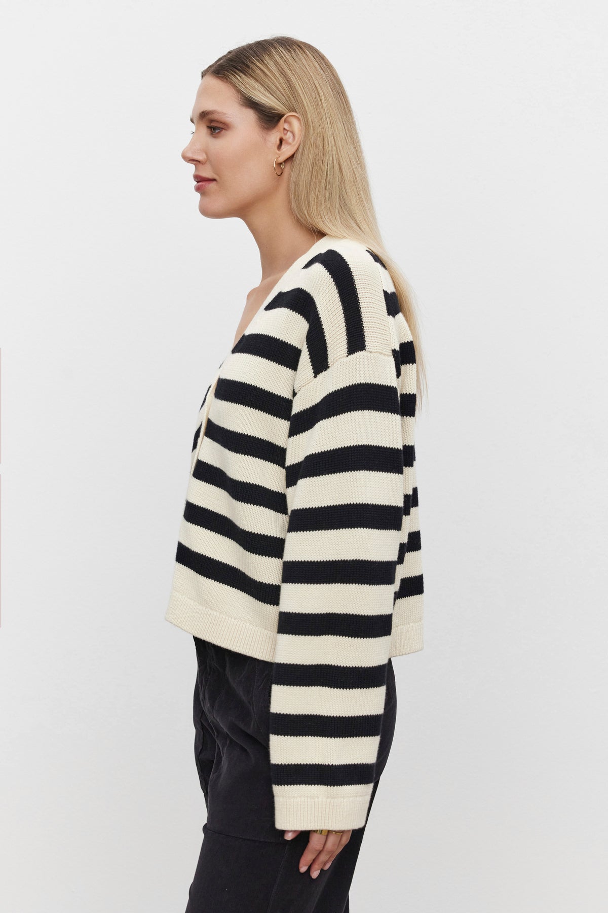   A woman with long blonde hair is wearing the ELODIE CARDIGAN from Velvet by Graham & Spencer, styled in a black and white striped cotton cashmere blend, paired with black pants, standing sideways against a plain white background. 