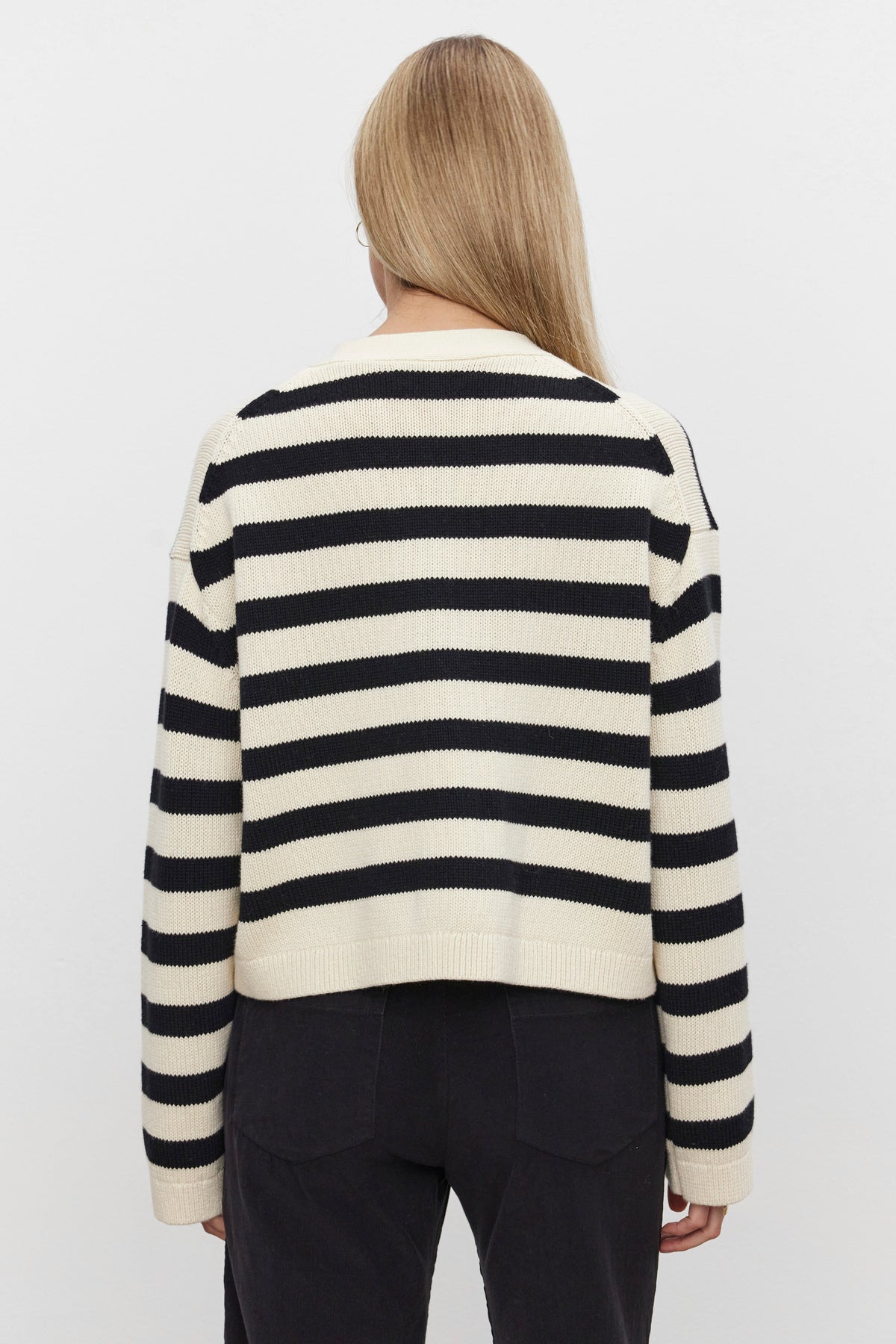   Person with long blonde hair wearing the ELODIE CARDIGAN by Velvet by Graham & Spencer, paired with black pants, facing away from the camera against a plain white background. 