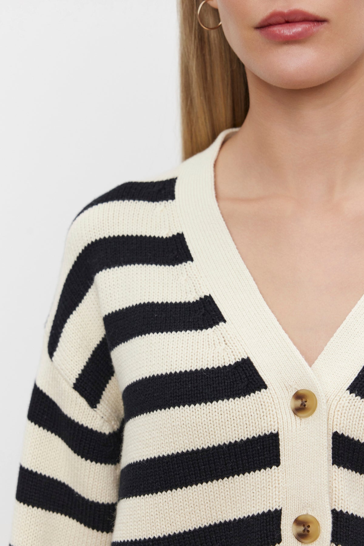   Close-up of a person wearing the ELODIE CARDIGAN by Velvet by Graham & Spencer, featuring a black and white striped pattern made from a cotton cashmere blend, three visible beige buttons, and a light-colored collar. One gold hoop earring is partially visible, set against a plain white background. 