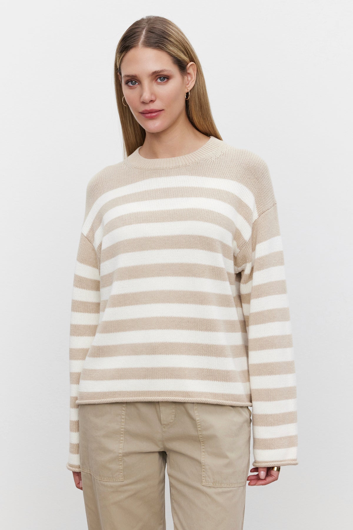   A person with long hair stands facing the camera, wearing the LEX SWEATER by Velvet by Graham & Spencer, made of a soft cotton cashmere blend and beige pants against a plain white background. 