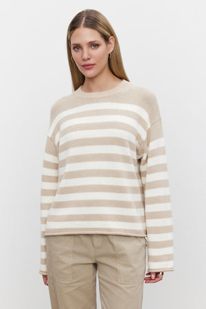 A person with long hair stands facing the camera, wearing the LEX SWEATER by Velvet by Graham & Spencer, made of a soft cotton cashmere blend and beige pants against a plain white background.