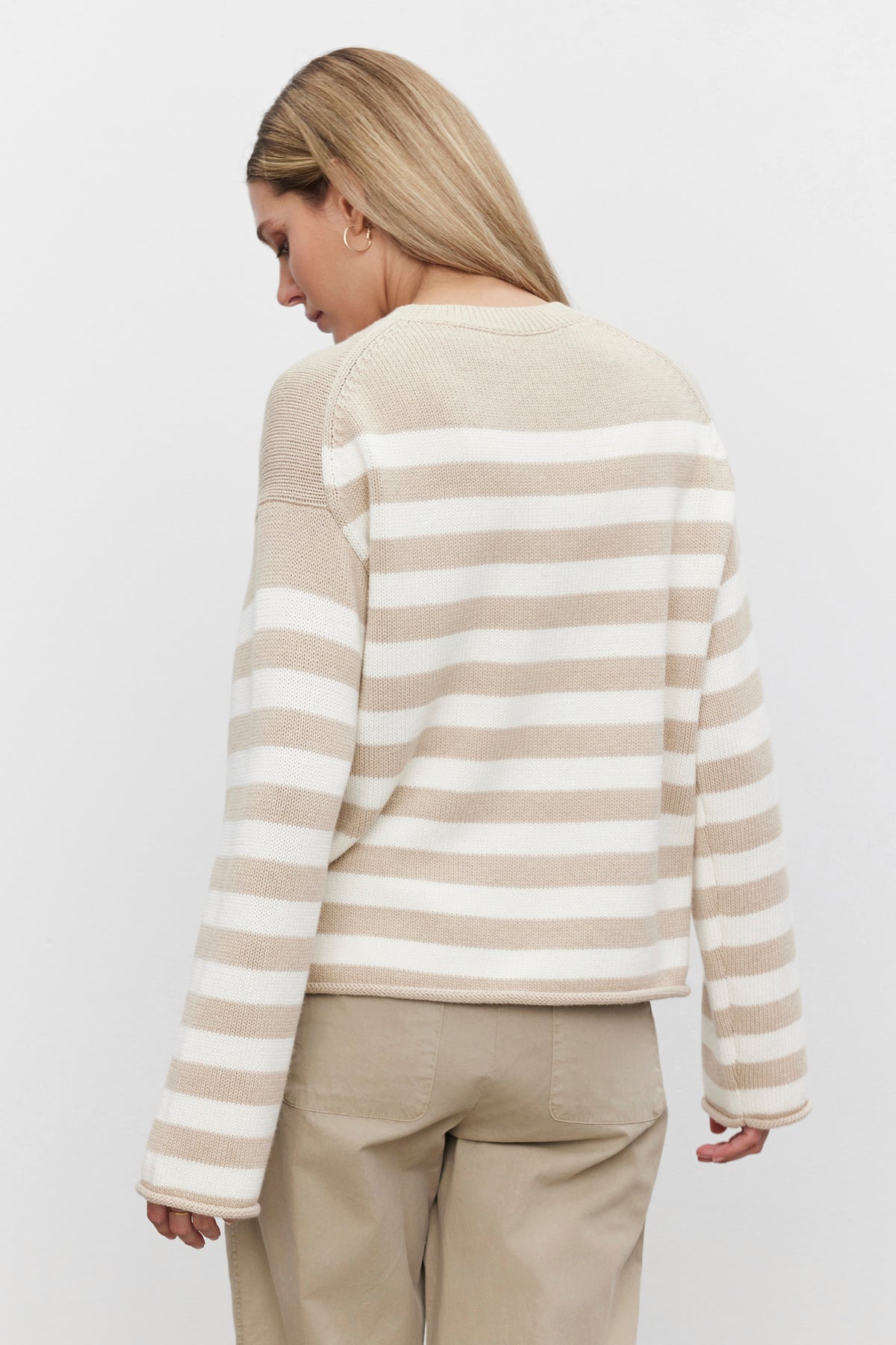   A person with long hair is facing away, wearing the LEX SWEATER by Velvet by Graham & Spencer, featuring modern beige and white stripes, paired with khaki pants. 