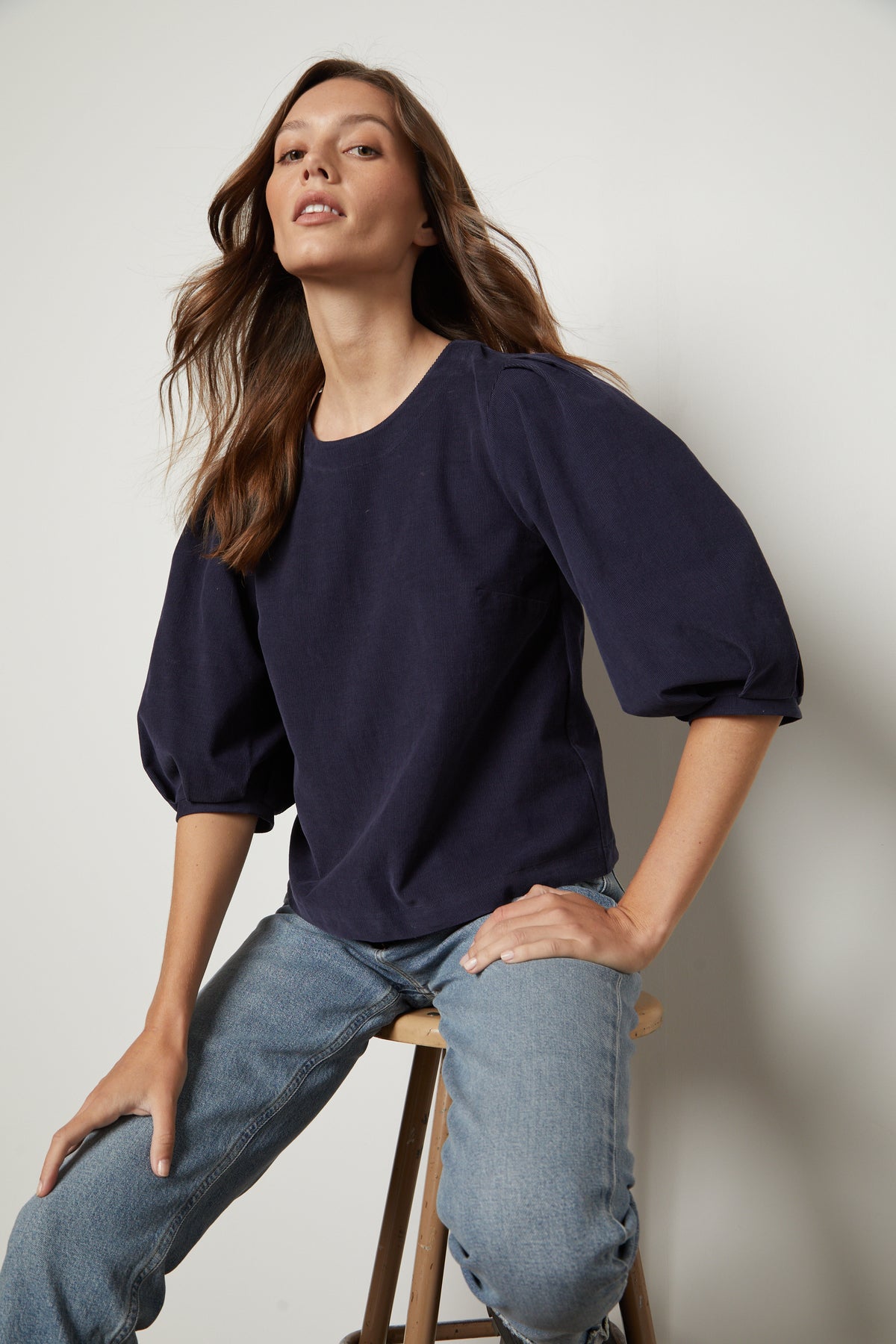   A woman is sitting on a stool wearing jeans and a navy Velvet by Graham & Spencer TARAH CORDUROY TOP with puff sleeves. 