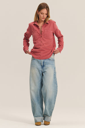 A woman stands against a plain background wearing the Velvet by Graham & Spencer ARIELLE CORDUROY SHIRT in pink and wide-leg cuffed light blue jeans, paired with classic brown shoes—perfect for updating your fall closet.