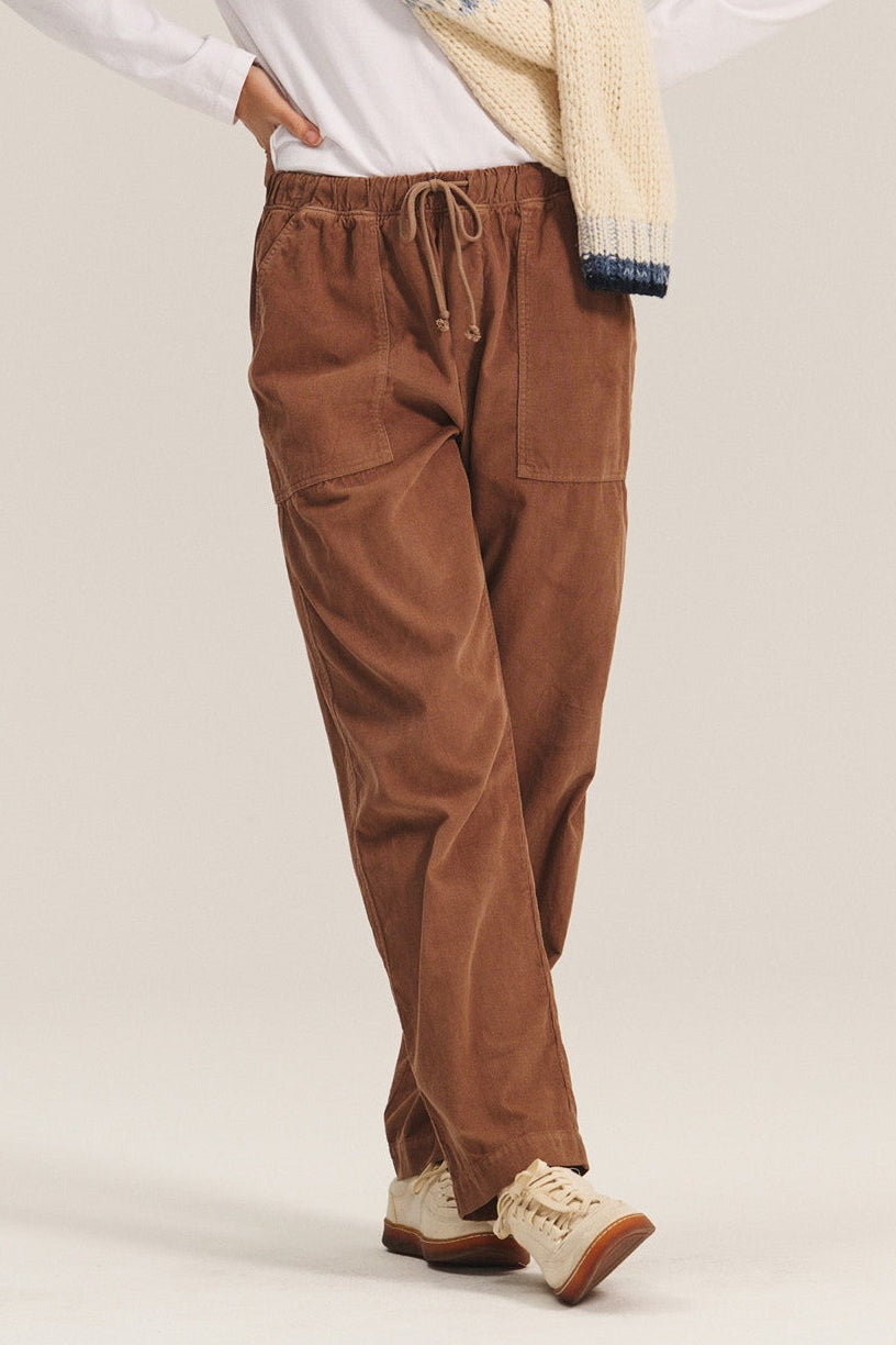   A woman stands with one hand on her hip, wearing Velvet by Graham & Spencer’s ASPEN CORDUROY PANT, a white long-sleeve shirt, beige sneakers, and a patterned knit sweater draped over her shoulders against a plain background. 