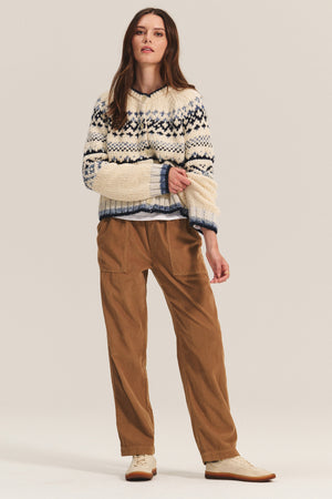 A woman wears the ASPEN CORDUROY PANT by Velvet by Graham & Spencer—brown corduroy pants with an elastic waist and large pockets—paired with a cream and navy patterned sweater and beige lace-up shoes against a plain backdrop.