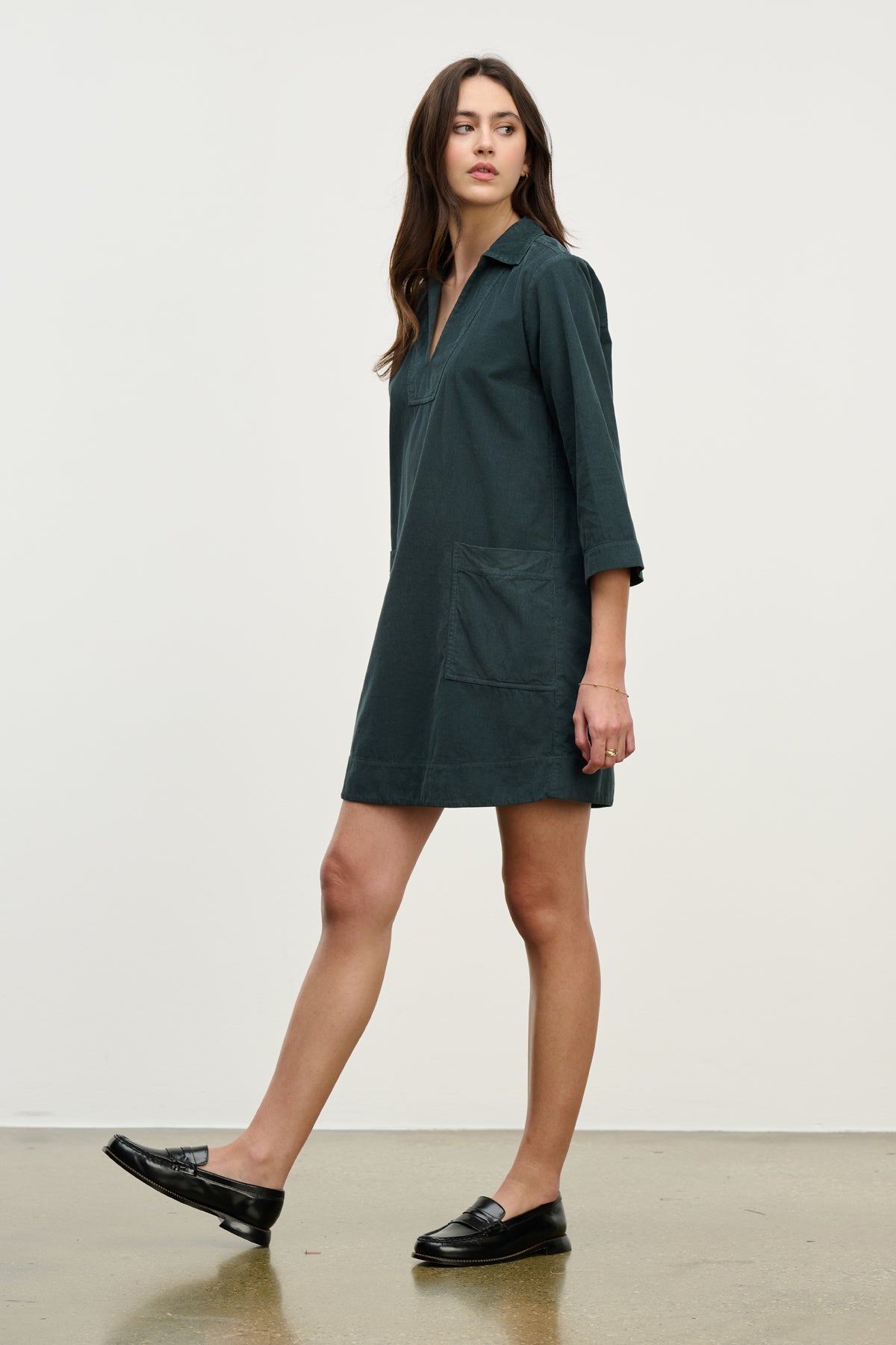 A woman in the ELKE CORDUROY DRESS by Velvet by Graham & Spencer—dark green, long-sleeve cotton with front pockets—pairs it with black loafers on a white backdrop, making it perfect for everyday wear.-39725334626497