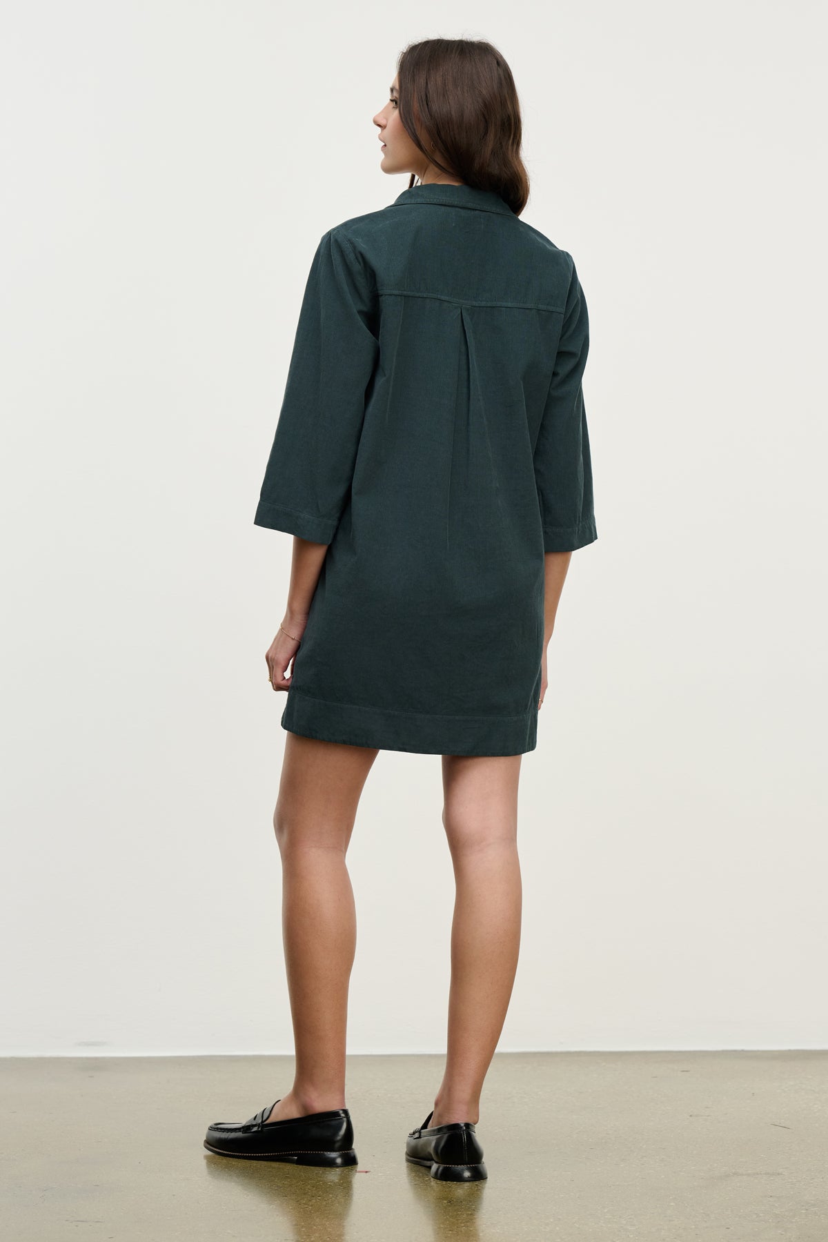 A woman faces away in the ELKE CORDUROY DRESS by Velvet by Graham & Spencer, a dark green cotton mini dress with three-quarter sleeves, paired with black loafers against a plain background.-39725334593729
