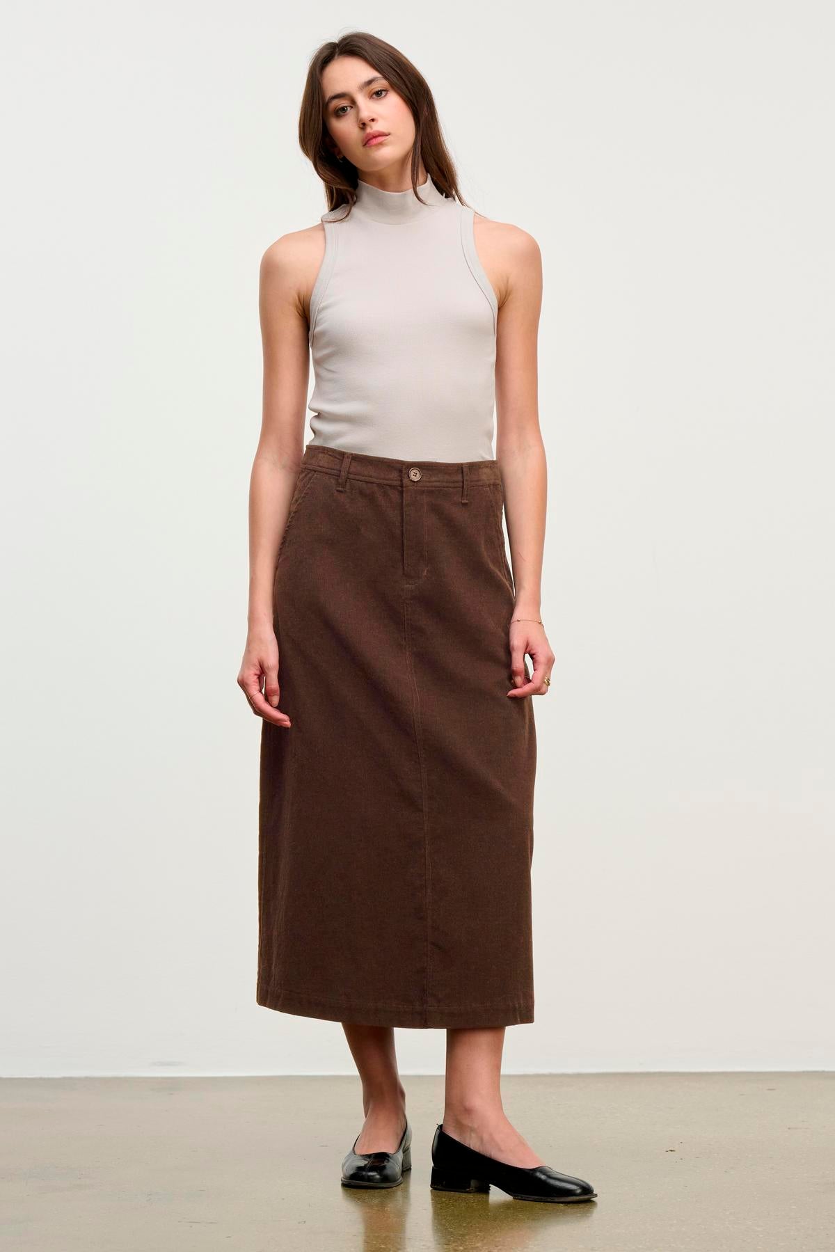 A woman stands against a plain background wearing a sleeveless beige turtleneck top, the FIA CORDUROY SKIRT by Velvet by Graham & Spencer in brown, and black flats.-39653622776001