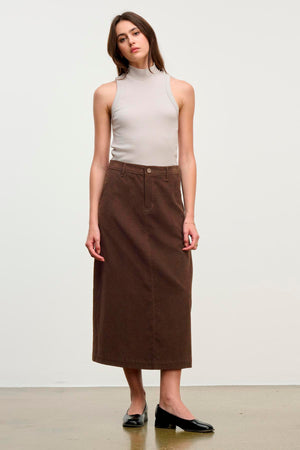 A woman stands against a plain background wearing a sleeveless beige turtleneck top, the FIA CORDUROY SKIRT by Velvet by Graham & Spencer in brown, and black flats.