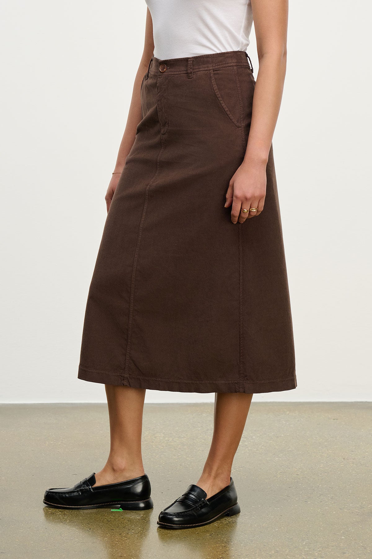 A person wearing a white sleeveless top, the FIA CORDUROY SKIRT by Velvet by Graham & Spencer featuring pockets and a button-zipper closure, and black loafers stands on a speckled floor.-39655053721793