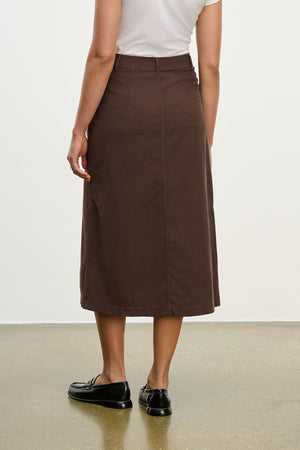 A person wearing a white short-sleeve shirt, black loafers, and the FIA CORDUROY SKIRT by Velvet by Graham & Spencer stands on a polished floor facing away from the camera.