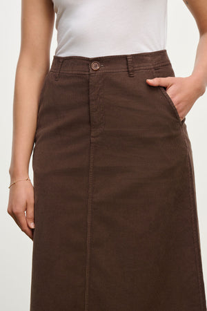 A person wears a white sleeveless top and the Velvet by Graham & Spencer FIA CORDUROY SKIRT—brown cotton with button and zipper closure—one hand in a pocket.