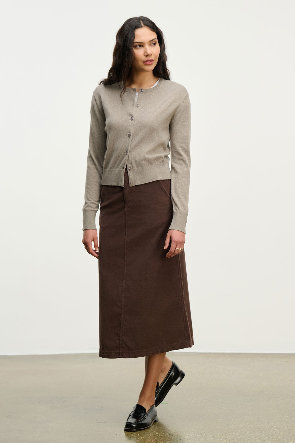   A person stands in a neutral space wearing the VIOLETTE CARDIGAN by Velvet by Graham & Spencer, a brown ankle-length skirt, and black loafers. 