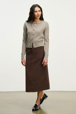 A person stands in a neutral space wearing the VIOLETTE CARDIGAN by Velvet by Graham & Spencer, a brown ankle-length skirt, and black loafers.