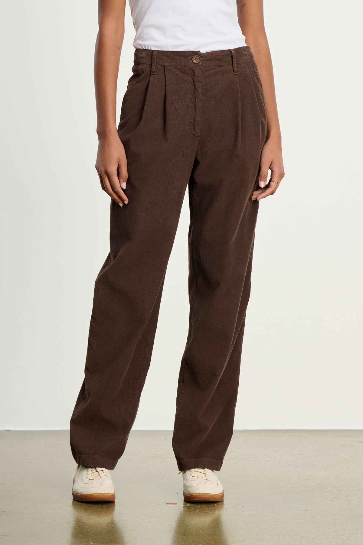 A person stands on a polished floor against a plain background, wearing Velvet by Graham & Spencer’s GAIL CORDUROY PANT in brown, paired with a white sleeveless top and light-colored sneakers.-39617449132225