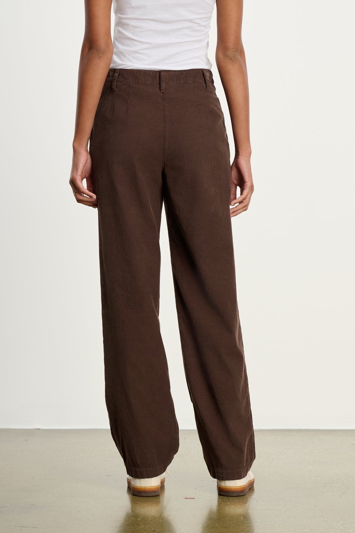 A person stands with their back to the camera, wearing a white sleeveless top and Velvet by Graham & Spencer's GAIL CORDUROY PANT in brown, styled with flat shoes.-39617449033921