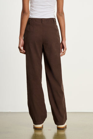 A person stands with their back to the camera, wearing a white sleeveless top and Velvet by Graham & Spencer's GAIL CORDUROY PANT in brown, styled with flat shoes.