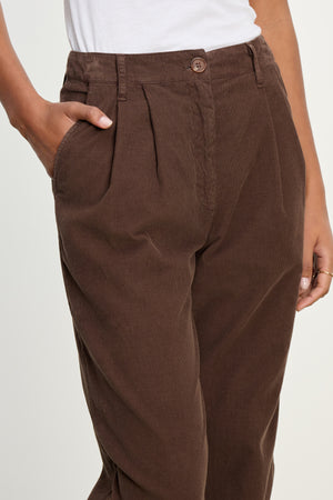 A person wearing Velvet by Graham & Spencer's GAIL CORDUROY PANT in brown with a white shirt, one hand in a pocket, shown from waist to thigh.