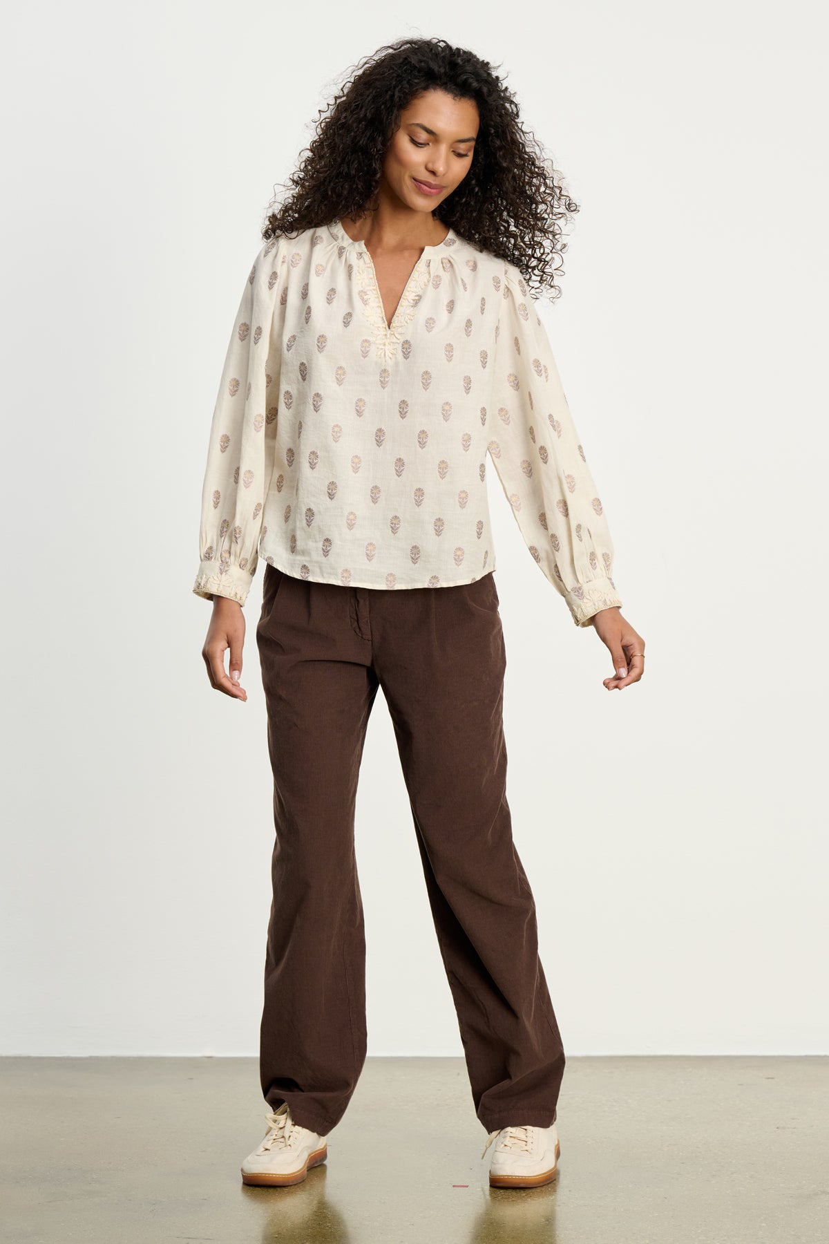 A woman stands indoors wearing a cream patterned blouse, light sneakers, and Velvet by Graham & Spencer's GAIL CORDUROY PANT in a straight-leg style against a plain background.-39617448902849