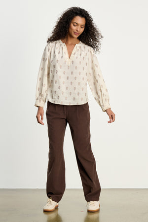 A woman stands indoors wearing a cream patterned blouse, light sneakers, and Velvet by Graham & Spencer's GAIL CORDUROY PANT in a straight-leg style against a plain background.