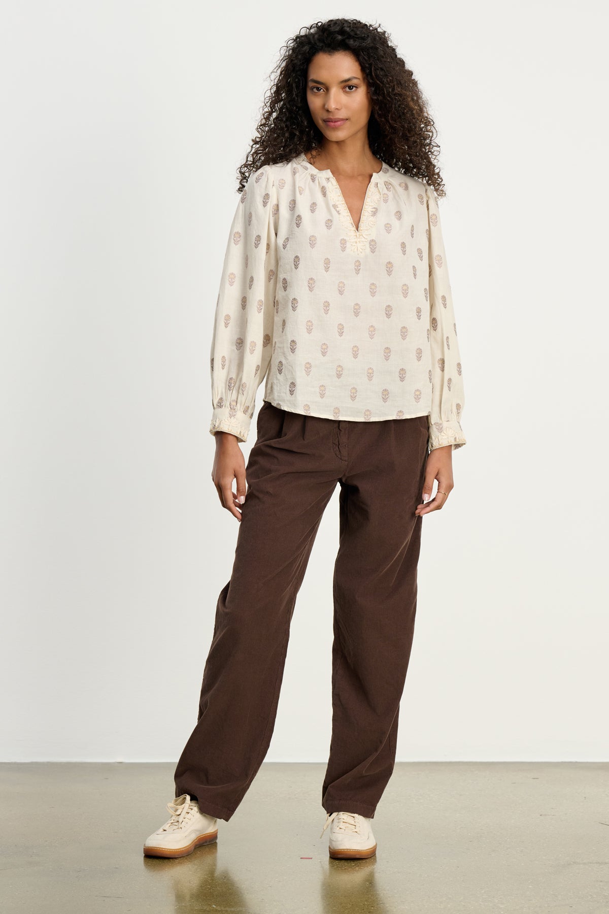 A woman stands against a white background wearing the GAIL CORDUROY PANT by Velvet by Graham & Spencer, paired with a beige patterned blouse and light-colored sneakers.-39617448837313
