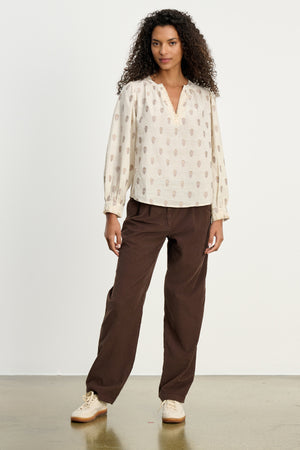 A woman stands against a white background wearing the GAIL CORDUROY PANT by Velvet by Graham & Spencer, paired with a beige patterned blouse and light-colored sneakers.