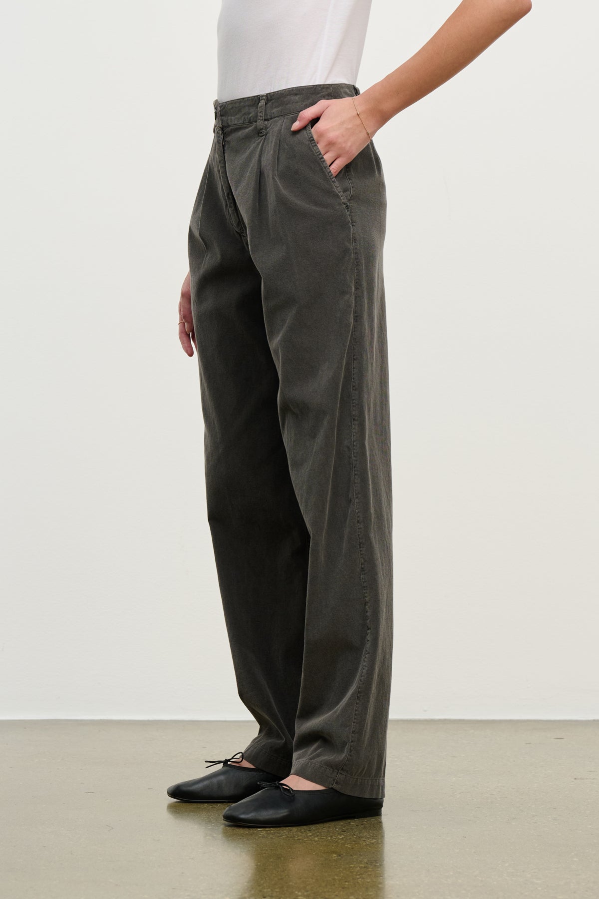 Someone wearing the GAIL CORDUROY PANT by Velvet by Graham & Spencer in loose-fitting dark gray stands against a plain background, paired with a white top and black ballet flats.-39800318132417