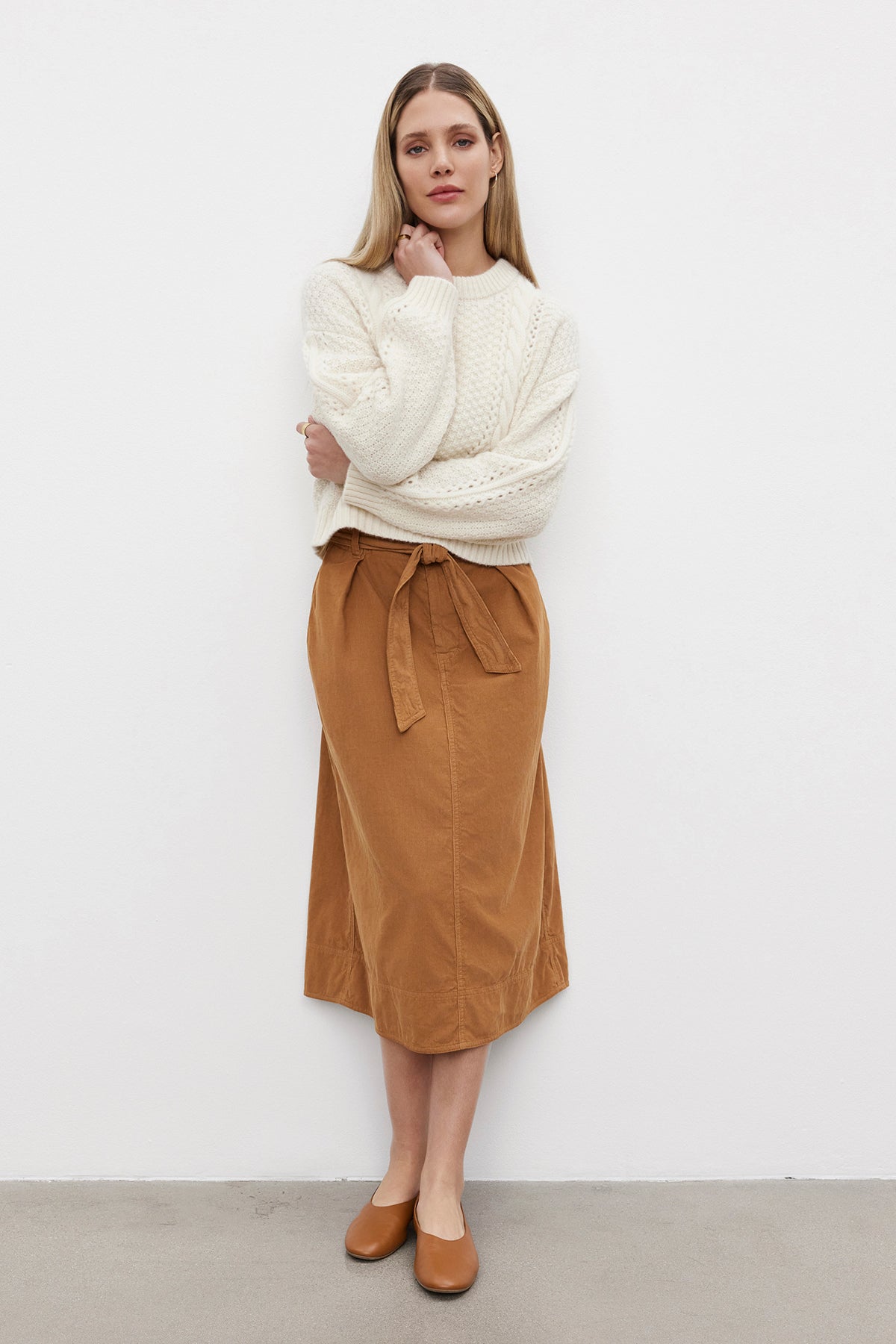   A woman stands against a white wall, wearing the MARLEY SWEATER by Velvet by Graham & Spencer—a cream-colored, cropped length alpaca blend sweater—paired with a tan midi skirt with a tied waist and matching tan shoes. She has long straight hair and is looking at the camera. 