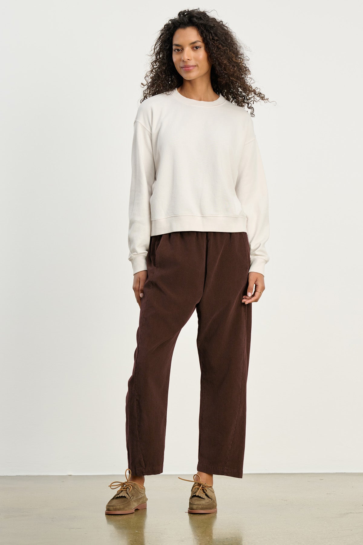 A person stands against a plain background wearing a white sweatshirt, tan lace-up shoes, and the LANEY CORDUROY PANT by Velvet by Graham & Spencer—cropped pinwale corduroy pants with an elastic waist.-40435578306753