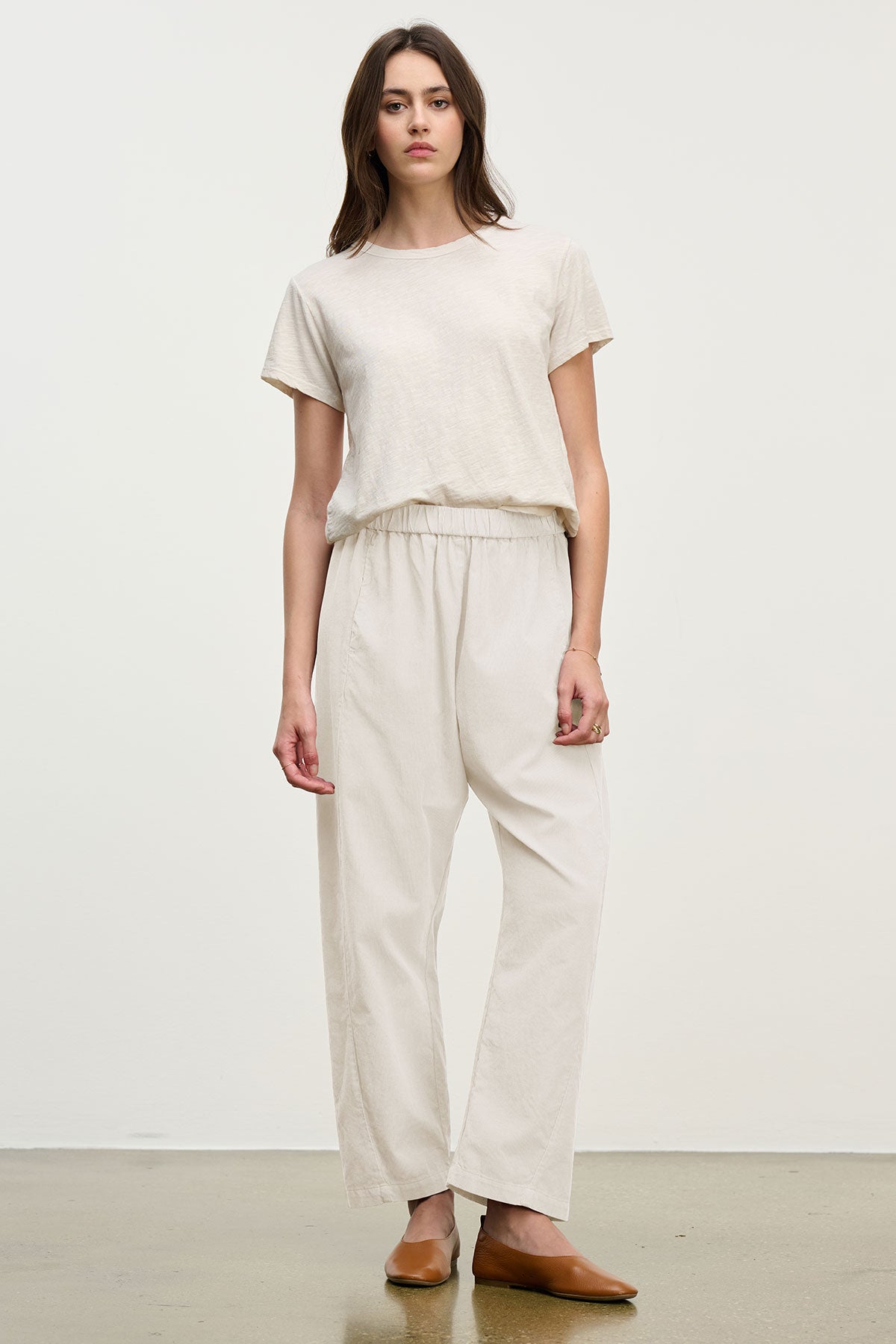 A person stands against a plain background wearing a beige t-shirt, brown flat shoes, and the LANEY CORDUROY PANT by Velvet by Graham & Spencer—cropped pinwale corduroy pants with an elastic pull-on waist.-39815566065857