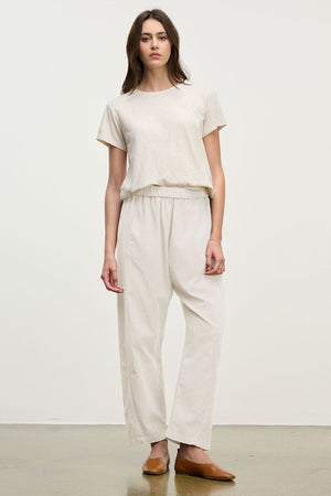 A person stands against a plain background wearing a beige t-shirt, brown flat shoes, and the LANEY CORDUROY PANT by Velvet by Graham & Spencer—cropped pinwale corduroy pants with an elastic pull-on waist.