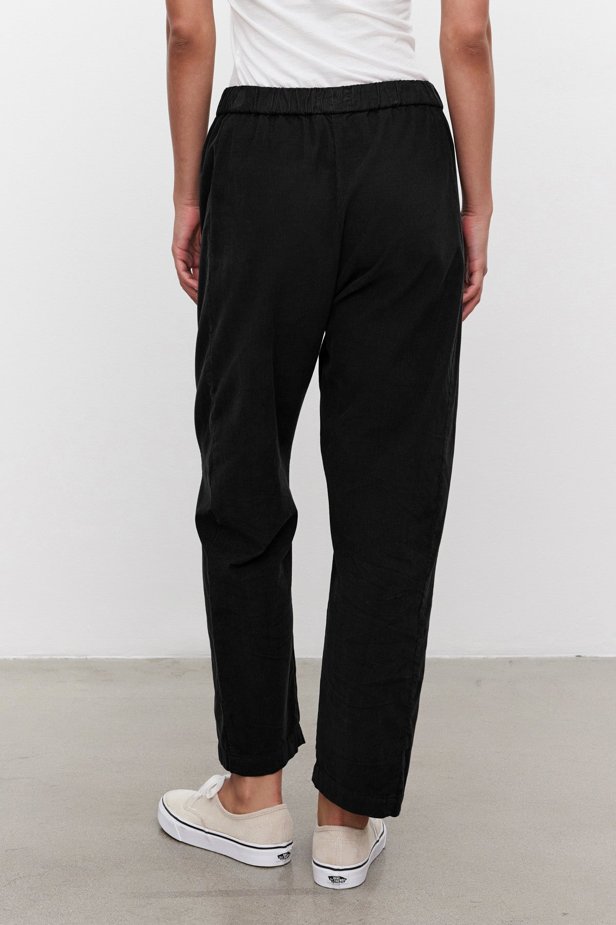 A person is seen from the back indoors, wearing the LANEY CORDUROY PANT by Velvet by Graham & Spencer in black pinwale corduroy with an elastic waist, styled with a white shirt and white slip-on sneakers on a light floor.