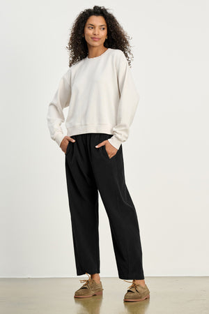 A person stands indoors against a plain background wearing a white sweatshirt, brown shoes, and the LANEY CORDUROY PANT by Velvet by Graham & Spencer, with hands in pockets.
