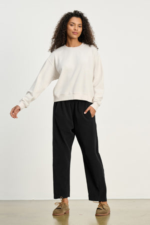A person stands against a plain background wearing a light-colored sweatshirt, brown shoes, and LANEY CORDUROY PANT by Velvet by Graham & Spencer.