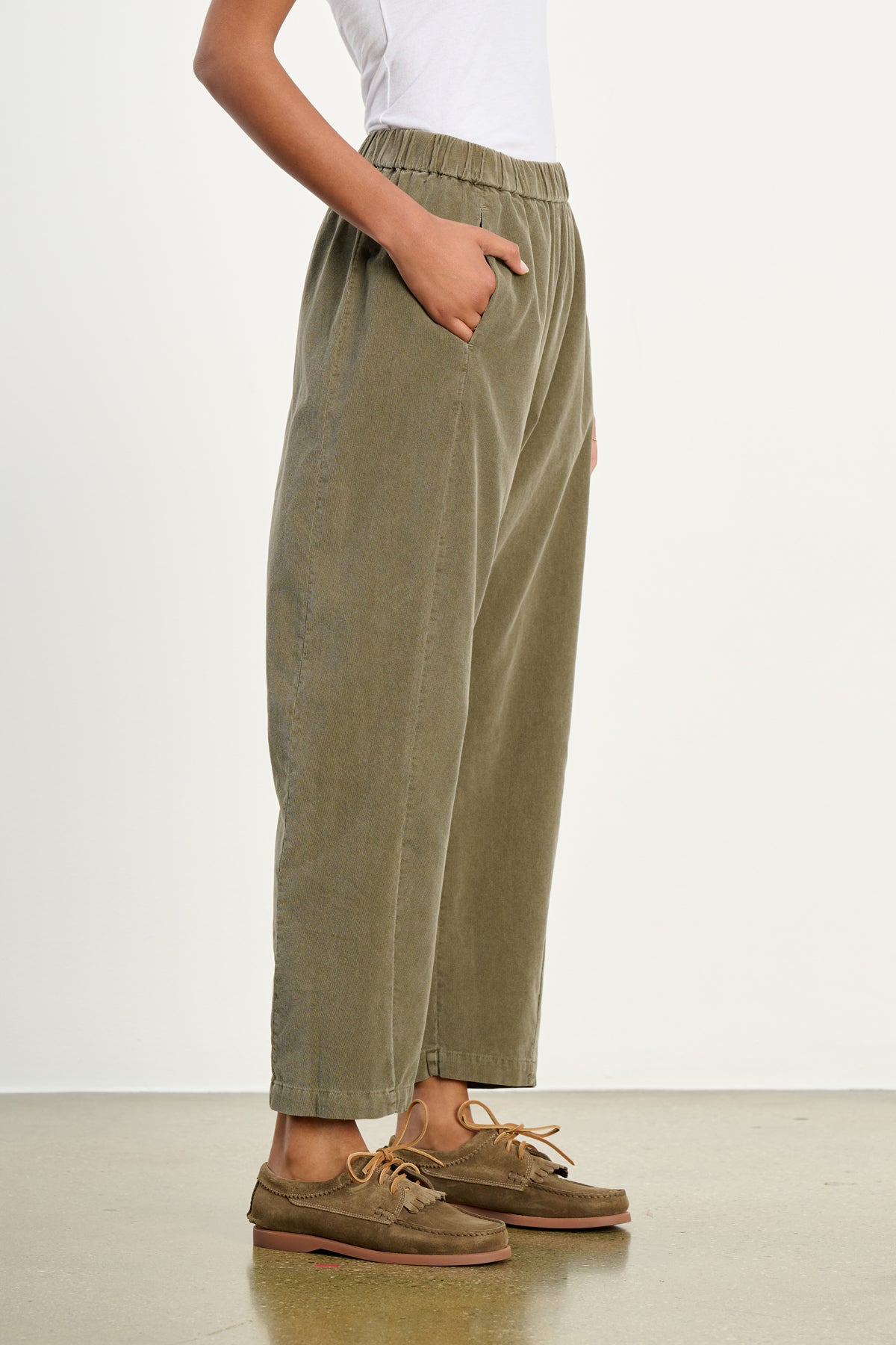 A person stands with hands in pockets wearing Velvet by Graham & Spencer's LANEY CORDUROY PANT in olive green, featuring a cropped fit and elastic pull-on waist, paired with a white top and brown boat shoes against a plain background.-39539208061121