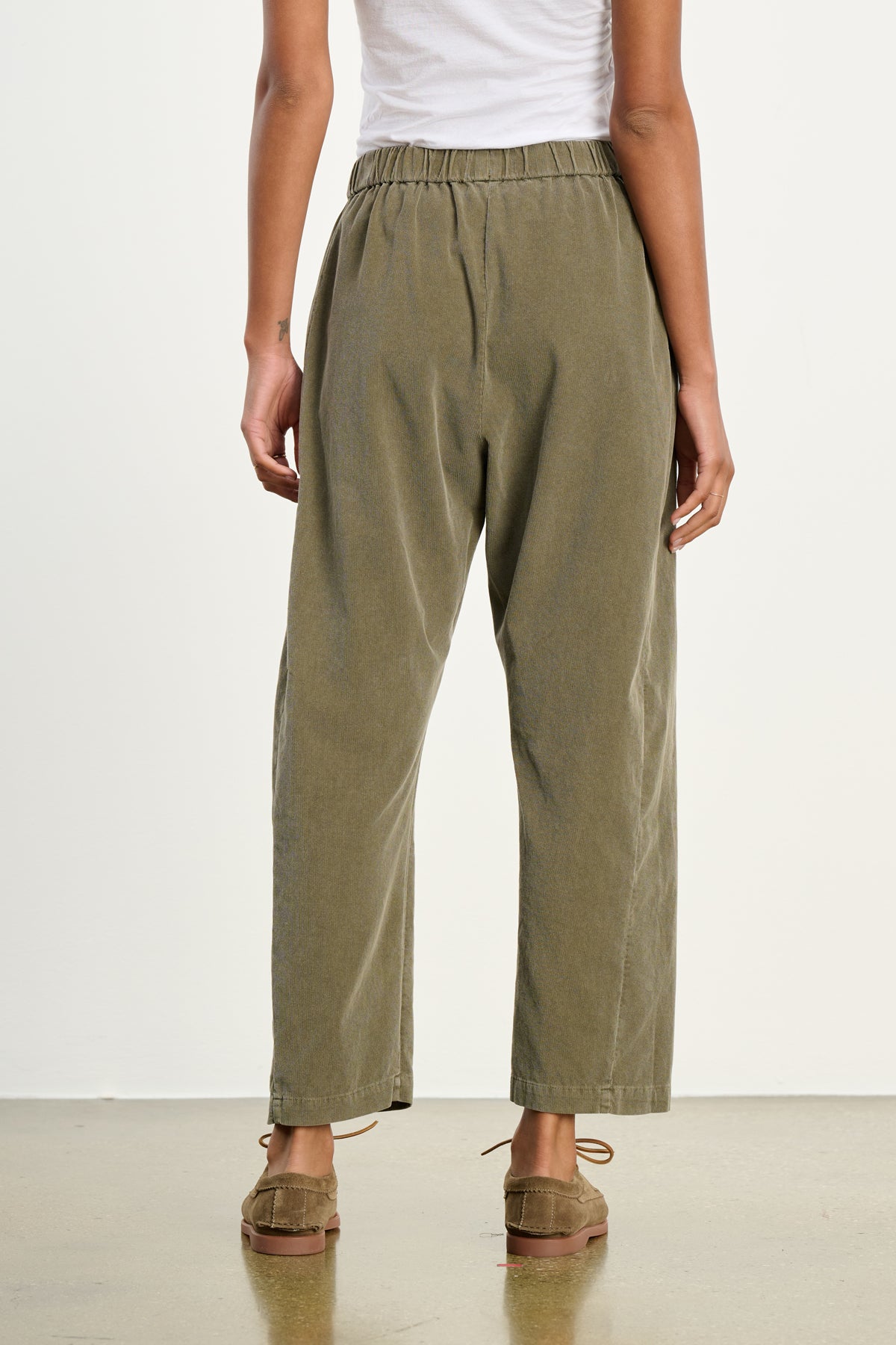 Person wearing Velvet by Graham & Spencer's LANEY CORDUROY PANT in olive green, featuring a loose fit and elastic pull-on waist, styled with a white top and brown moccasins, shown from the back on a polished floor against a plain white background.-39539208028353