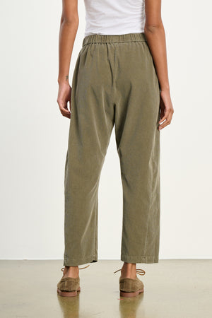 Person wearing Velvet by Graham & Spencer's LANEY CORDUROY PANT in olive green, featuring a loose fit and elastic pull-on waist, styled with a white top and brown moccasins, shown from the back on a polished floor against a plain white background.