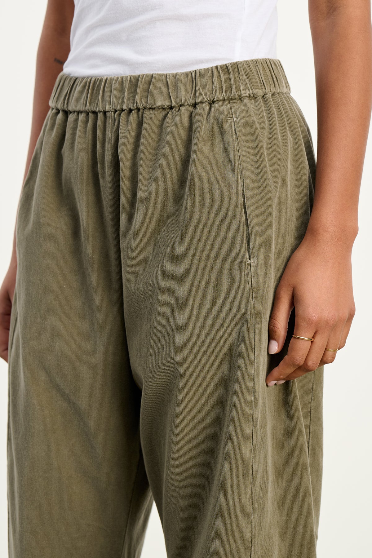 A person stands against a plain background wearing Velvet by Graham & Spencer's LANEY CORDUROY PANT in olive green, featuring pinwale corduroy and an elastic drawstring waist, paired with a white top. One hand rests by their side.-39539207995585