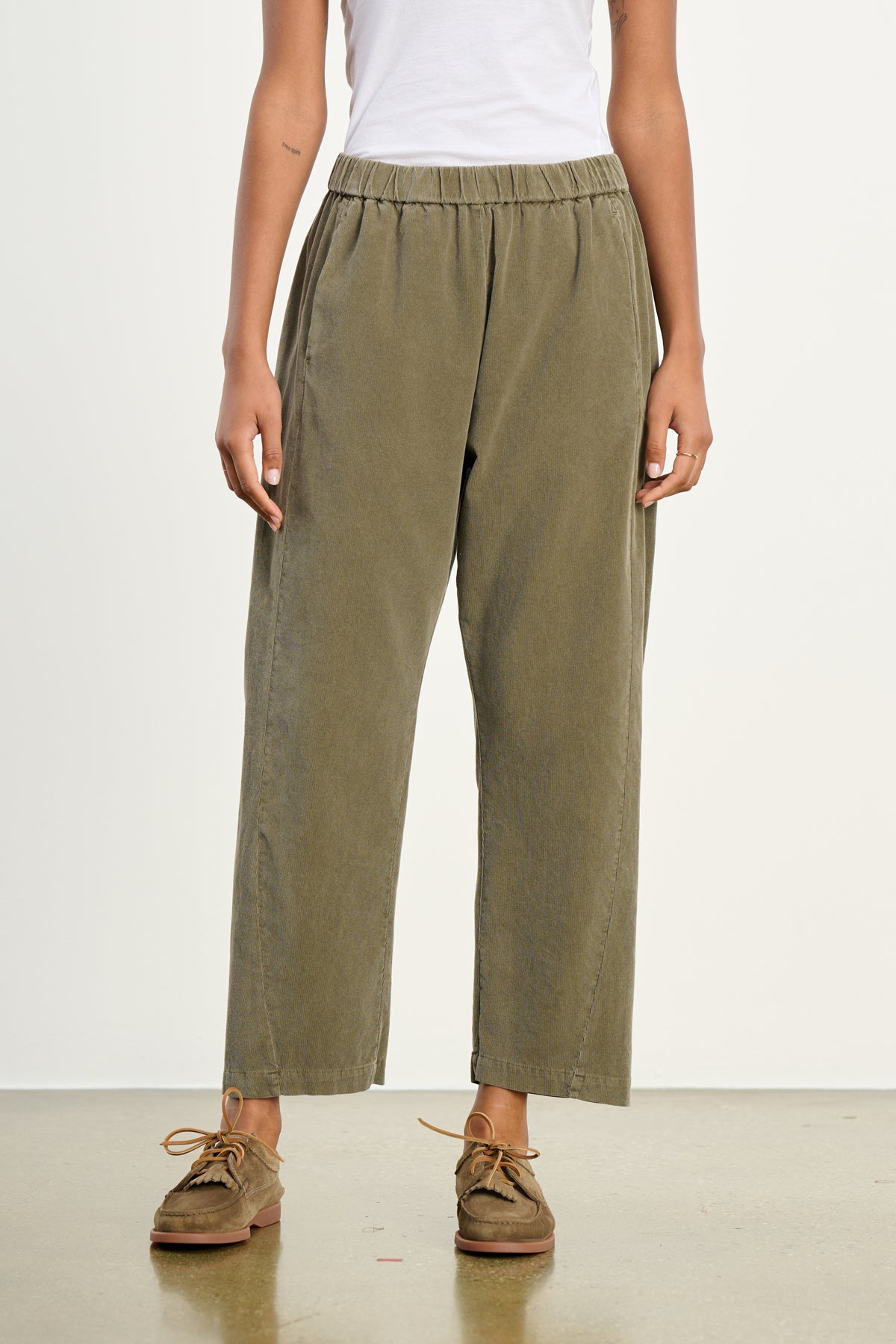 A person wears the LANEY CORDUROY PANT by Velvet by Graham & Spencer—olive green, high-waisted, cropped pinwale corduroy pants with an elastic pull-on waist—paired with a white sleeveless top and brown suede shoes against a plain background.-39539207962817