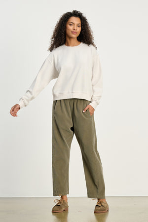 A person stands against a plain background wearing a light-colored sweatshirt, brown shoes, and the LANEY CORDUROY PANT by Velvet by Graham & Spencer—an olive green, elastic pull-on waist pinwale corduroy cropped pant.