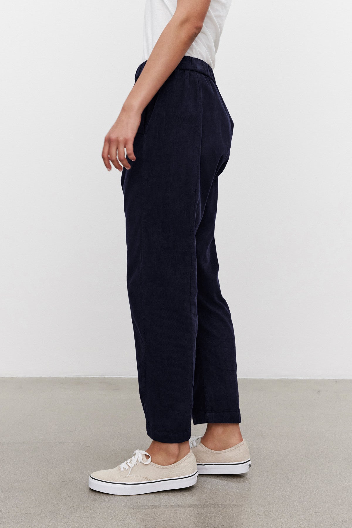 A person stands sideways on a light background wearing a white shirt, beige sneakers, and the LANEY CORDUROY PANT by Velvet by Graham & Spencer in navy blue with an elastic waist.