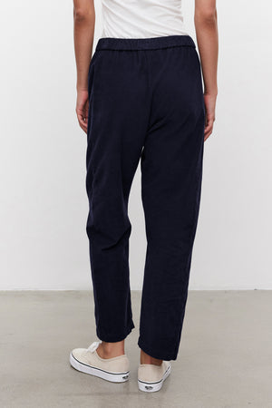 A person stands indoors, facing away, wearing Velvet by Graham & Spencer's LANEY CORDUROY PANT in dark navy with a relaxed, cropped fit and elastic waist, paired with a white top and white slip-on sneakers.