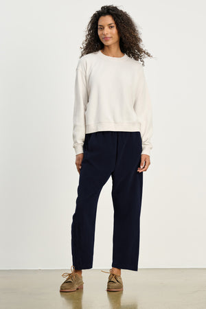 A person stands against a plain background wearing a white sweatshirt, brown shoes, and the LANEY CORDUROY PANT by Velvet by Graham & Spencer—cropped pinwale corduroy pants with an elastic waist.