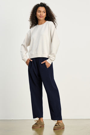 A woman stands against a plain background wearing a cream sweatshirt and Velvet by Graham & Spencer’s LANEY CORDUROY PANT in navy blue with an elastic waist, her hands tucked in her pockets, and brown shoes.