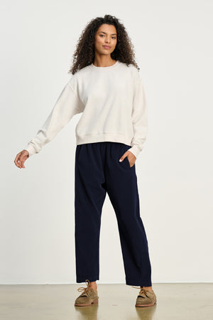 A person stands in a neutral pose, wearing a cream sweatshirt and the LANEY CORDUROY PANT by Velvet by Graham & Spencer in navy with an elastic waist, paired with brown shoes against a plain background.