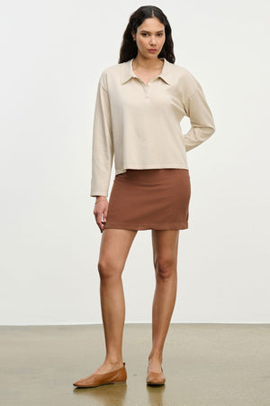 A woman stands against a plain background wearing a beige long-sleeve polo shirt, brown flats, and the LIZA CORDUROY SKIRT by Velvet by Graham & Spencer.