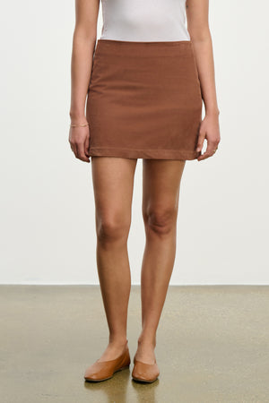 A person in a white sleeveless top, the LIZA CORDUROY SKIRT by Velvet by Graham & Spencer—a fitted cotton mini skirt—and brown flats stands on a polished floor against a plain white background.